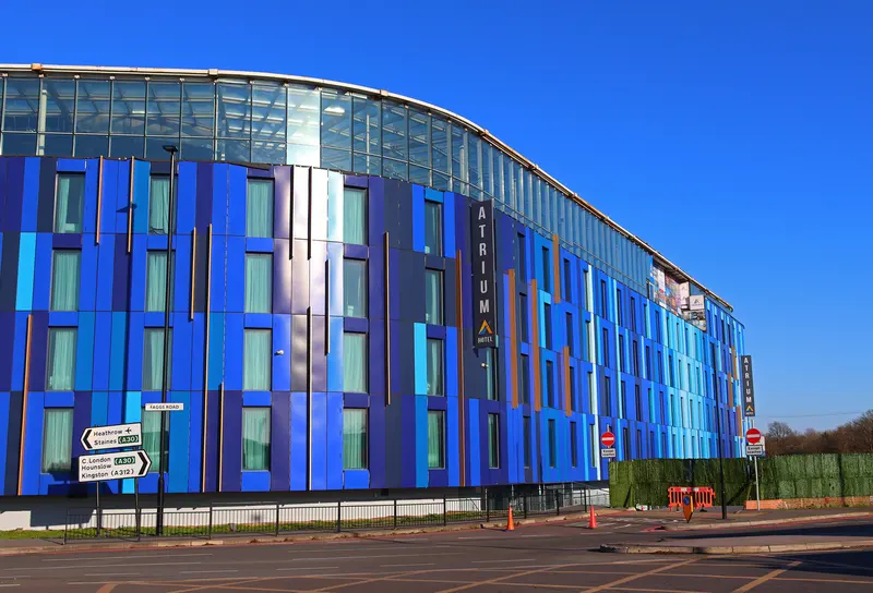 A curved hotel clad in blue panels with a glass top floor and a vertical ATRIUM HOTEL sign, beside road signs to Heathrow and Staines.