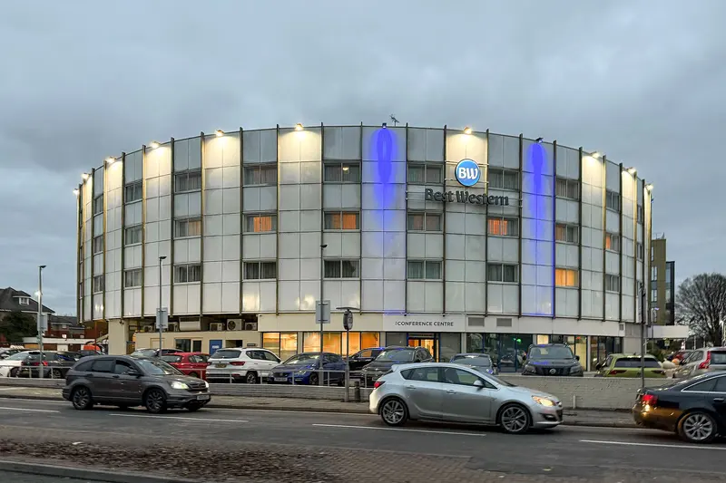 A circular white hotel building lit at dusk with a Best Western sign and logo on its front, beside the Bath Road.